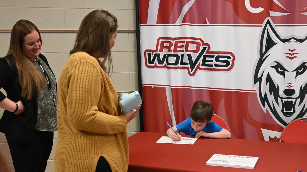 principals standing with student seated in front of red wolves backdrop